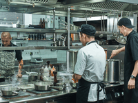 The image shows three chefs actively working in a busy, modern commercial kitchen. The atmosphere is focused and professional, typical of a restaurant or high-volume food production setting.