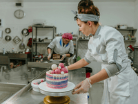 In the center of the image, a woman is holding a beautifully decorated cake on a stand. She's wearing a white chef's coat and disposable gloves. Her hair is styled in a high bun, and she has a striped headband on. The cake she's holding has striped frosting in shades of purple, pink, and white, and is decorated with light pink and white spheres.