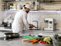 A chef wearing a white toque and uniform is working in a brightly lit, modern kitchen. He is standing at a stainless steel sink, washing some orange vegetables