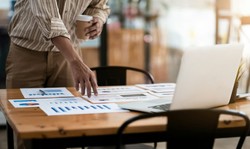 Man analyzing graphs on papers on the table, with a laptop and a cup of coffee beside him