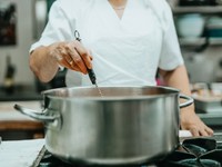 The image shows a chef in a professional kitchen stirring a large stainless steel pot on a stovetop.