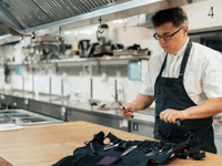 The image shows a chef standing in a professional kitchen, sharpening a knife with a honing steel. He is wearing glasses, a white chef’s jacket, and a dark apron.