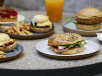 The image showcases a variety of well-presented gourmet dishes — including a deli-style bagel, eggs benedict, artisan burger, fries, and fresh juice — designed to convey quality, variety, and professionalism. The clean, polished setup reflects the brand’s ability to cater to corporate events, hotels, cafes, and businesses looking for reliable food service partners.