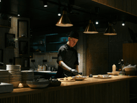 This is a photograph of a chef working at the pass or service counter of a kitchen with a modern, dark design aesthetic.