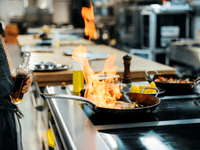 The image captures a close-up, dynamic moment of flambé cooking in a professional kitchen environment.