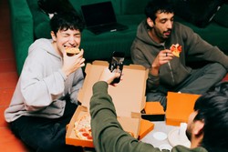 Friends enjoying a casual meal from a food delivery service, gathered around a green couch with pizza boxes and slices of pizza in hand. One person captures the moment by taking a photo with their smartphone.