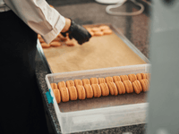 This close-up photo shows someone's hands, wearing black gloves, working on a large batch of macaron cookies in a kitchen.