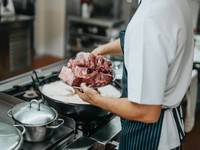 The image shows a chef in a professional kitchen holding a plate filled with raw chunks of red meat, possibly beef. The chef is standing in front of a large pot on a stove, which appears to be filled with a boiling liquid—likely preparing for a stew, broth, or soup.