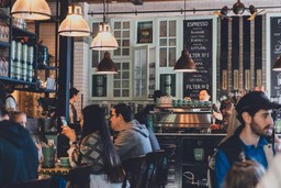 Customers enjoying coffee and meals in a cozy, well-lit cafe with a barista preparing drinks in the background.
