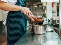 The image shows a chef in a professional kitchen serving food from a stainless steel pot into a small dish using a ladle. The chef is wearing a striped apron, indicating a food service or culinary professional.