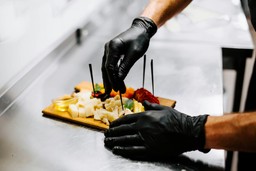 A chef wearing black gloves preparing a charcuterie board with various cheeses, meats, and garnishes on a stainless steel counter.