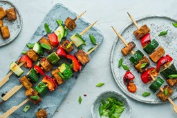 Skewers with grilled tofu, zucchini, and red bell peppers garnished with sliced green onions on stone and ceramic plates.
