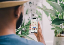 A man holding a smartphone with a restaurant map and location markers on the screen, possibly searching for nearby dining options using a food delivery or restaurant discovery app. Surroundings include plants and a casual setting.