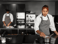 This photograph features a female chef actively working in a modern, professional kitchen, with a colleague preparing food in the background.