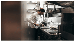 A professional chef wearing a white jacket stands in a modern commercial kitchen, carefully chopping ingredients on a cutting board.