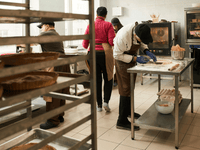 The image shows a busy commercial bakery kitchen with several people actively engaged in baking tasks. In the foreground, a person in a white chef's coat, brown apron, and black cap is working at a stainless steel table, using a fork on a sheet of dough.