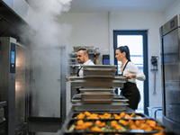In a professional kitchen, two chefs (a man and a woman) are seen working together. Both are wearing white shirts and dark aprons. The woman is holding a tray, while the man is standing near a commercial oven, which is releasing a large amount of steam into the room.